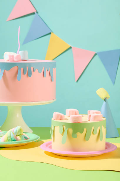 istock Front view of two birthday cakes made from colored paper decorated with marshmallows. A triangular flag string hangs on a blue background. Copy space for birthday. 1957043769