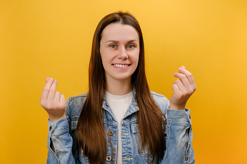 Portrait of attractive smiling young woman 25s looking at camera with money or italian gesture with hands, wearing denim jacket, posing isolated over plain yellow color background wall in studio