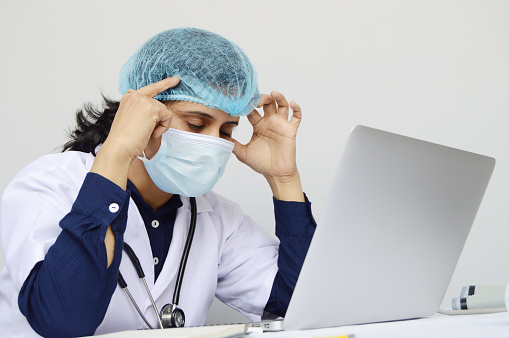 Horizontal photo of one stressful woman nurse or doctor or health care worker, physician in headache tired while working on laptop over gray background with copy space for text.