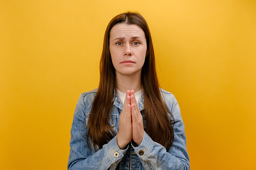 Portrait of young woman holding hands in prayer and asking for help with pleading imploring expression, hope concept, wearing denim jacket, isolated over yellow studio background. Please, I need