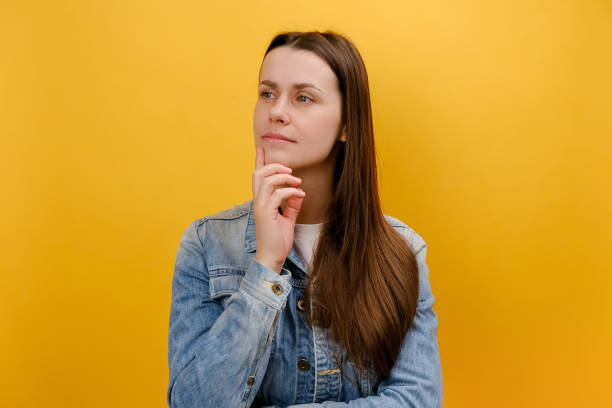 istock Portrait of pensive young woman holding her chin and seriously thinking over problem solution, having thoughtful expression, wearing denim jacket, isolated over yellow color background wall in studio 1957150363