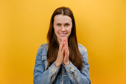 Portrait of tricky cunning smiling young caucasian woman standing looking at camera, planning something, pranking somebody, wearing denim jacket, posing isolated over yellow background wall in studio