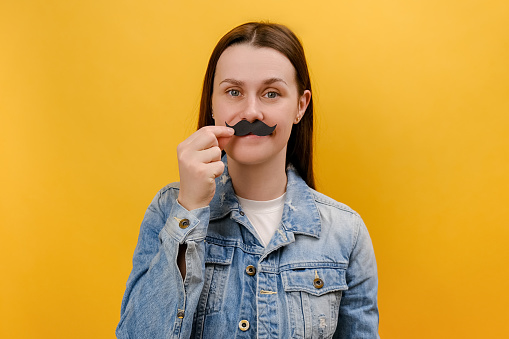 Portrait of funny playful young woman keeping fake paper mustache, happy looking at camera, having festive mood, wearing denim jacket, posing isolated over plain yellow color background wall in studio