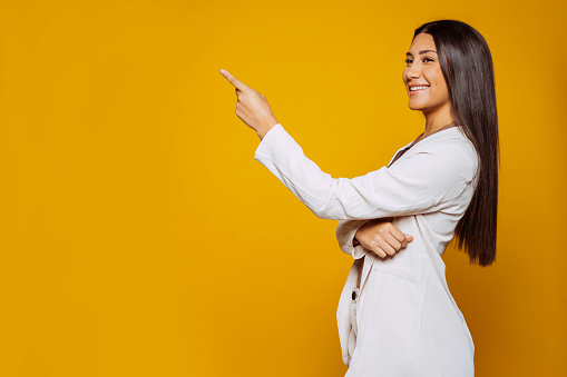 Portrait of beautiful smiling latin american businesswoman in suit, looking away, pointing to the side at studio isolated over yellow background.