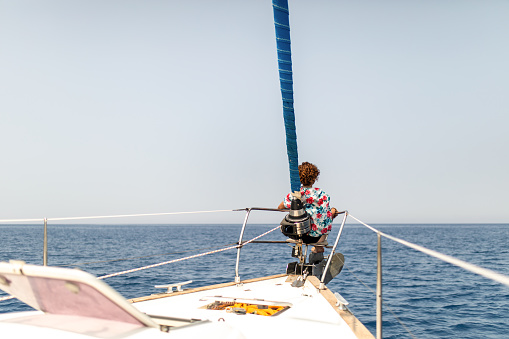 Mature woman sitting on a sailboat bow, relaxing and looking towards the horizon over sea. She is alone, carefree and pensive.