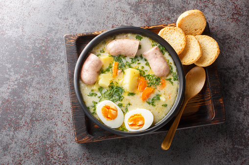Polish Sour Rye Soup Zurek with white sausage, boiled egg and fresh veggies close-up in a bowl on the table. Horizontal top view from above