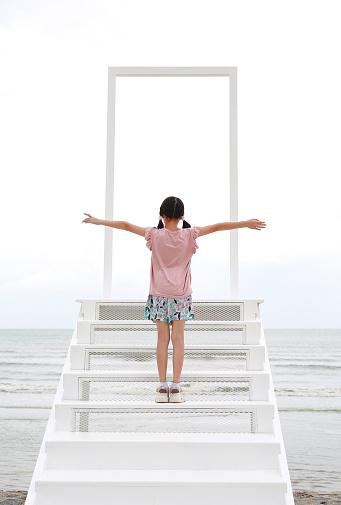 Back view of Asian girl child open hands outstretched or keeping arms raised. Kid standing on open door gate with stairs to the sea and sky.