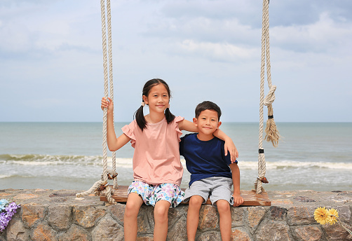 Adorable Asian little boy child and older girl kid sitting together on wooden swing against cement wall at seaside.