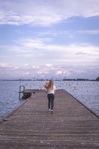 Carefree young woman running on pier, araound her birds are flying
