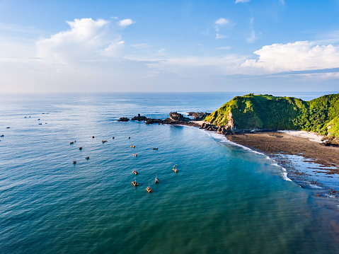 A lush green hillside meets the clear blue waters of the ocean. The rocky shoreline is visible, adorned with various shapes and sizes of rocks. In the distance, a cloudy sky paints a serene backdrop. Shot taken on Lombok.