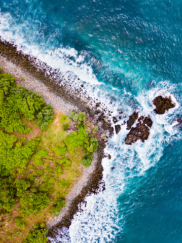 Aerial view capturing the vibrant contrast between the turquoise ocean waves crashing against a rocky shoreline and a green coastal landscape adorned with lush foliage.