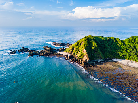 A lush green hillside meets the clear blue waters of the ocean. The rocky shoreline is visible, adorned with various shapes and sizes of rocks. In the distance, a cloudy sky paints a serene backdrop.