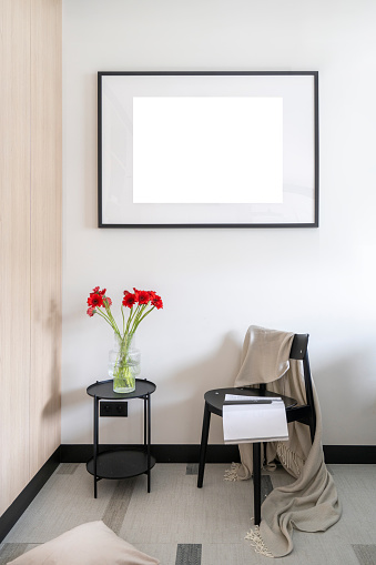 Vertical shot of coffee table with glass vase with flowers, black wooden chair with plaid against white wall with picture. Copy space. Cozy home interior.