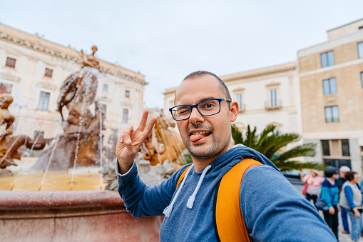 Mid-adult man taking selfies in front of the fountain of Diana in Syracuse on the Italian island of Ortygia.