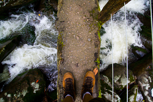 Journey into the Wild: A fearless adventurer crosses a weathered bridge over a rushing river, showcasing sturdy hiking boots against the untamed beauty of Gatlinburg, Tennessee. The perfect image for outdoor exploration and overcoming challenges.