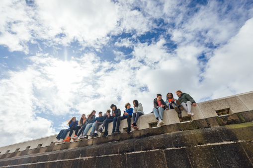Low angle wide view of a large group of teenage friends wearing casual clothing on a summer day in Whitley Bay, Northeastern England. They are gathered sitting along the wall on the promenade in a line side by side, taking selfies on smartphones and talking.