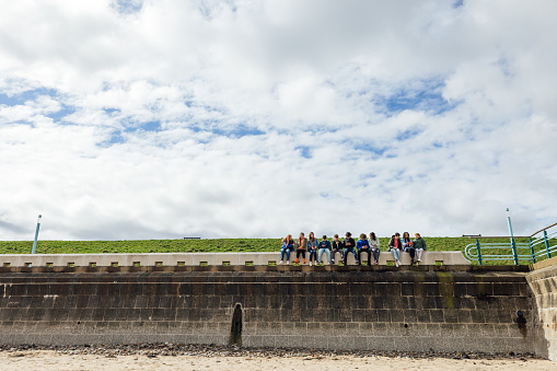 Wide front view of a large group of teenage friends wearing casual clothing on a summer day in Whitley Bay, Northeastern England. They are gathered sitting along the wall on the promenade in a line side by side, taking selfies on smartphones and talking.