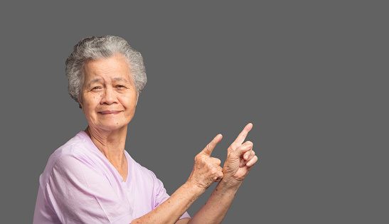 Portrait of an elderly Asian woman with short gray hair pointing with two hands and fingers to the side and looking at the camera with a smile while standing on a gray background. Space for text