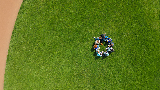 Directly above drone view of a large group of teenage friends wearing casual clothing on a summer day in Whitley Bay, Northeastern England. They are gathered sitting on the grass in a circle together talking and eating chips.
