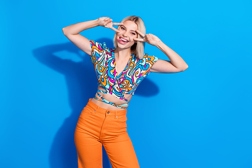 Photo of cute woman with bob hairdo dressed print blouse stick out tongue showing v-sign on eyes isolated on vibrant blue color background.