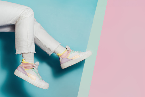 Close up female legs in white jeans and retro style high-top multicolor sport sneakers shoes on multicolor blue and pink background. Pastel candy colors, vintage retro style of 80s - 90s vibes