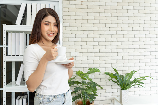 Portrait of a smiling young brunette woman dressed casually holding a white cup of hot coffee while sitting at a table at a cafe, natural lighting lifestyle photo.
