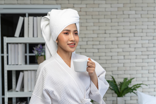 Cropped view of young woman wearing a white bathrobe and wet hair wrapped in a soft white towel, holding a coffee mug, standing in the bedroom