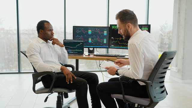 Two men traders sitting at desk at office together monitoring stocks data candle charts on screen