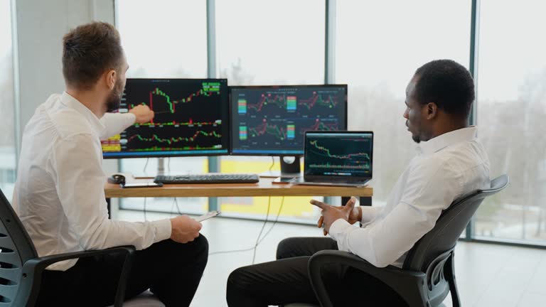 Two diverse colleagues traders talking to each other, looking at graphs while sitting in the office in front of multiple computer screens