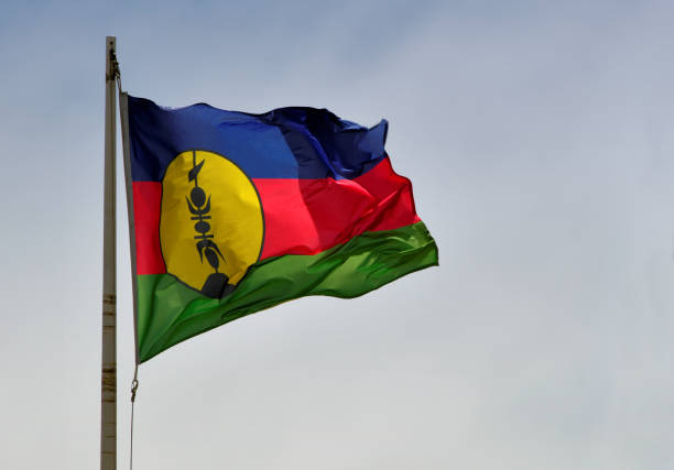 Kanak flag / Flag of New Caledonia (actual photo, not CGI) - 'fleche faitiére', Nouméa, New Caledonia Nouméa, South Province, New Caledonia:  Kanak flag / Flag of New Caledonia, indigenous Melanesian used in parallel with the French flag, originally the flag of the independence movement FLNKS. The Flag consists of three equal horizontal bands of blue (top), red, and green. A large yellow disk edged in black and displays a black 'fleche faitiére' (rooftop arrow). Emblem of the Kanak chiefdom, the 'fleche faitiére', is a traditional element of Kanak housing, it is a wood sculpture which dominates and adorns the cone-shaped thatched roof of the large huts and ceremonial huts of a clan. fleche stock pictures, royalty-free photos & images