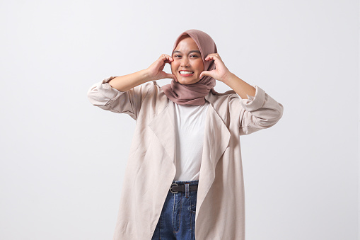 Portrait of attractive Asian hijab woman in casual suit speaks about own feelings, makes heart gesture over chest, expresses sympathy and love, smiles positively. Isolated image on white background