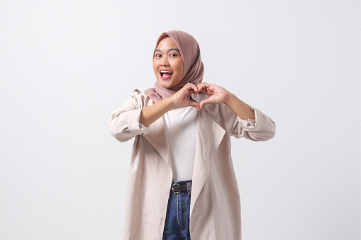 Portrait of attractive Asian hijab woman in casual suit speaks about own feelings, makes heart gesture over chest, expresses sympathy and love, smiles positively. Isolated image on white background