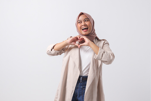Portrait of attractive Asian hijab woman in casual suit speaks about own feelings, makes heart gesture over chest, expresses sympathy and love, smiles positively. Isolated image on white background