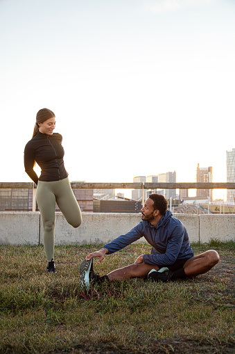 Vertical portrait of young multiracial couple stretching leg before running, training outdoors. Copy space. Fitness and relationships.