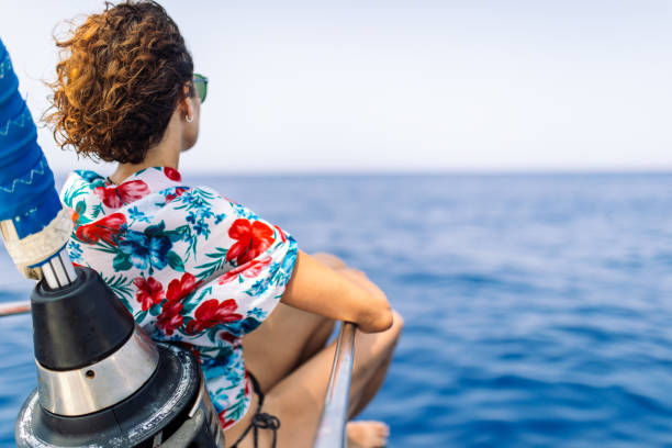 Mature woman sitting on a sailboat bow, relaxing and looking towards the horizon over sea. She is alone, carefree and pensive.