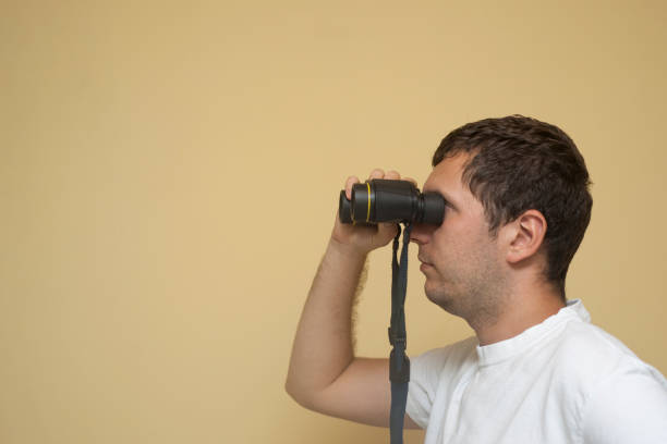 A young white man is viewing in binoculars far away on a neutral background.
