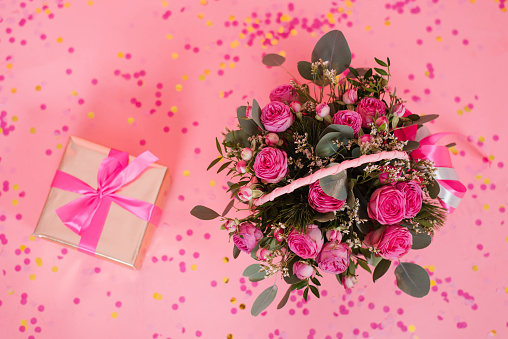 Bouquet of pink roses in a wicker basket and a gift box with a bow on a pink table background. Birthday, Wedding, Mother's Day, Valentine's Day, Women's Day. Top view