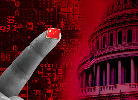Concept exploring the ever-present threat of China to American National Security.  A human finger holders a microchip with a Chinese flag on it against the backdrop of motherboard and the United States Capitol