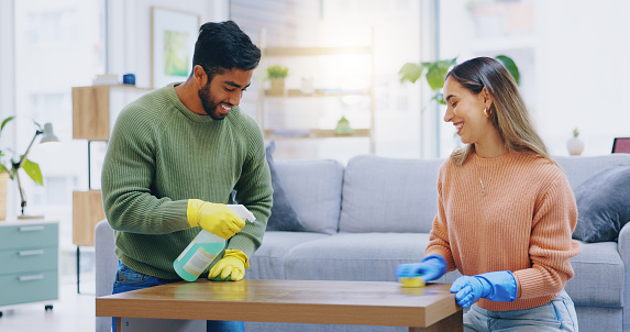 Couple, spray and cleaning in home of table, furniture and disinfection of dust, dirt or risk of bacteria. Happy man, interracial woman and bottle of chemical detergent to wipe surface in living room