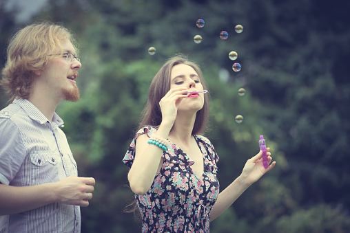 Happiness and carefree concept. Young woman and man having fun blowing soap bubbles together in park, green blurred background