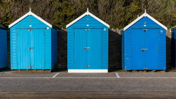 Three beach huits in different shades of blue, on a promenade in Dorset. Taken on a sunny December afternoon. Part of a series.