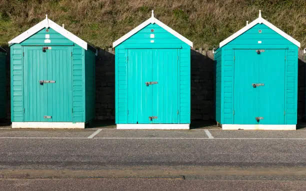 Three beach huts in shades of aqua blue on a promenade in Dorset. Taken on a sunny December afternoon. Part of a series.