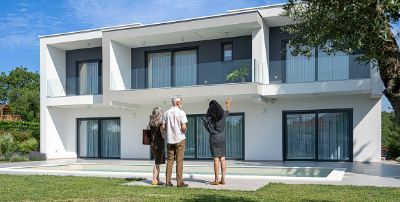 Female real estate agent showing modern luxury house to mature couple during sunny day.
