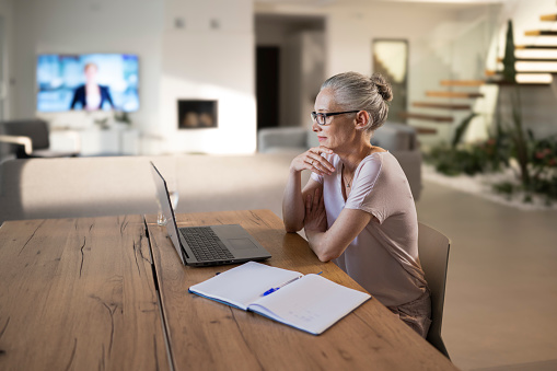 Mature businesswoman using laptop during conference call while sitting at table in living room.