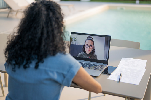 Businesswoman talking to female colleague during conference call on laptop besides swimming pool.
