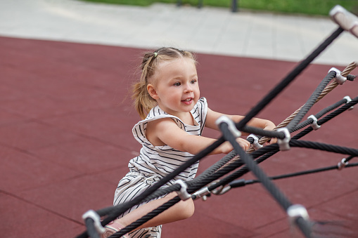 Toddler girl in striped outfit climbs with joy on rope playground equipment, with focus and determination.