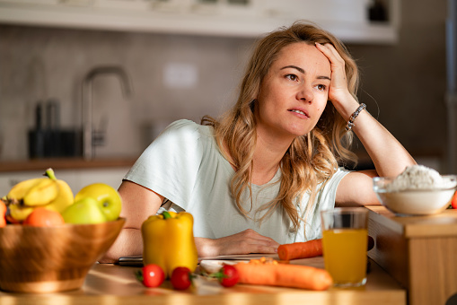 A tired woman is trying to prepare lunch while sitting in the kitchen and looking into the distance