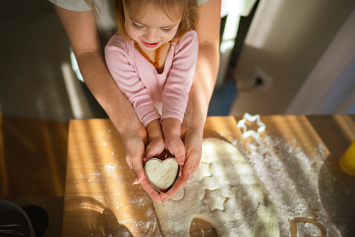 High angle view of a little girl holds the dough in the shape of a heart with the help of her older sister in the kitchen