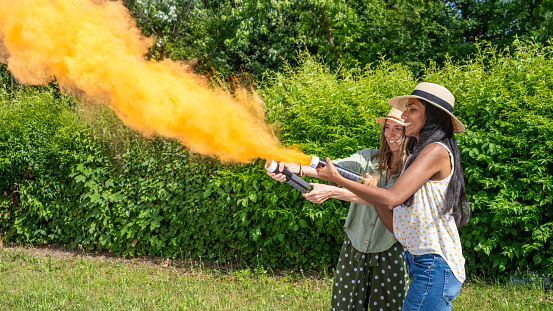 Two multiracial women shooting orange powder cannon outdoors at festive summer day, medium shot