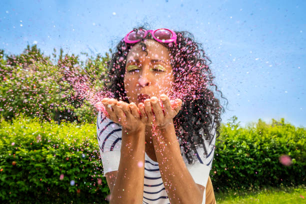 Mid adult hispanic woman blowing pink glitter from palms at camera outdoors at sunny day, medium shot